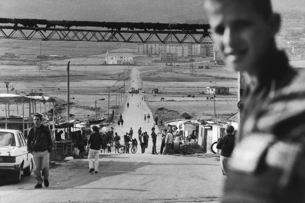 Bustling street market along a straight road; pedestrians, bicycles, stalls line the sides, a blurred person dominates the right foreground.