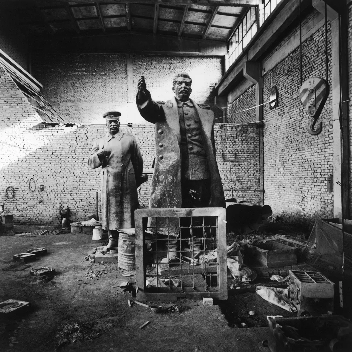 Statue of a man in a long coat raising his arm in an abandoned brick-walled factory; a second uniformed statue stands nearby.