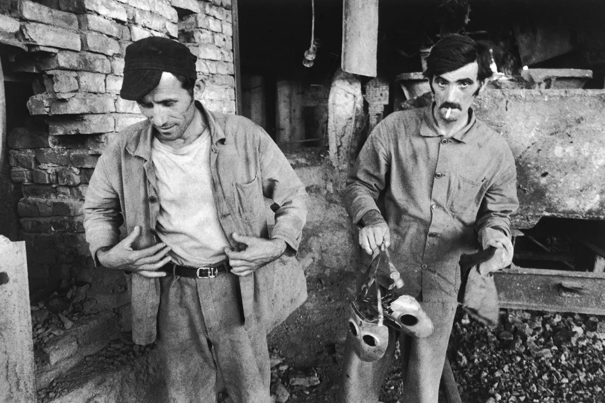 Two laborers in a rough brick workshop; left man opens his jacket, right man holds a gas mask and strap beside dusty machinery.