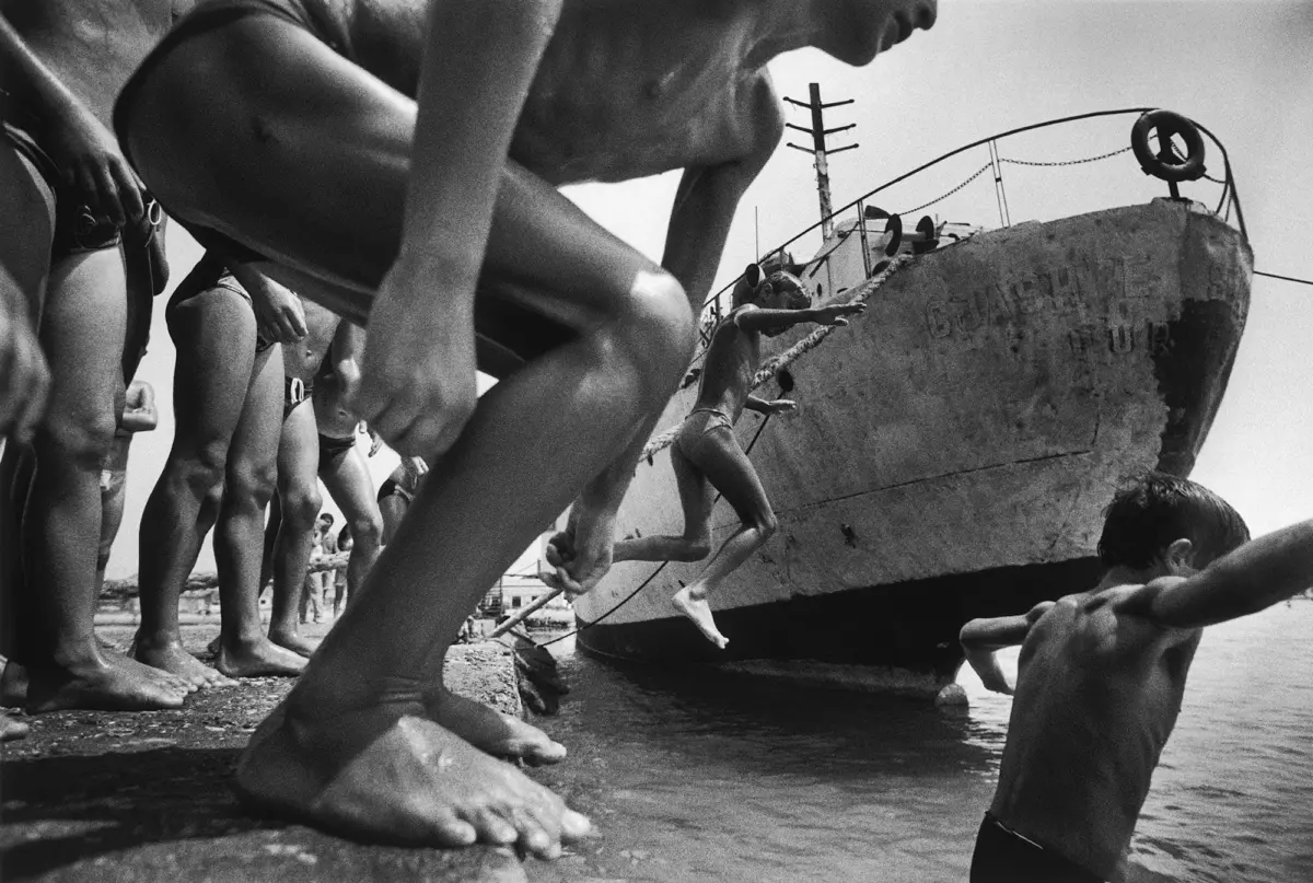 Group of barefoot people lined up on a beach, boarding a weathered boat with a lifebuoy on its bow.