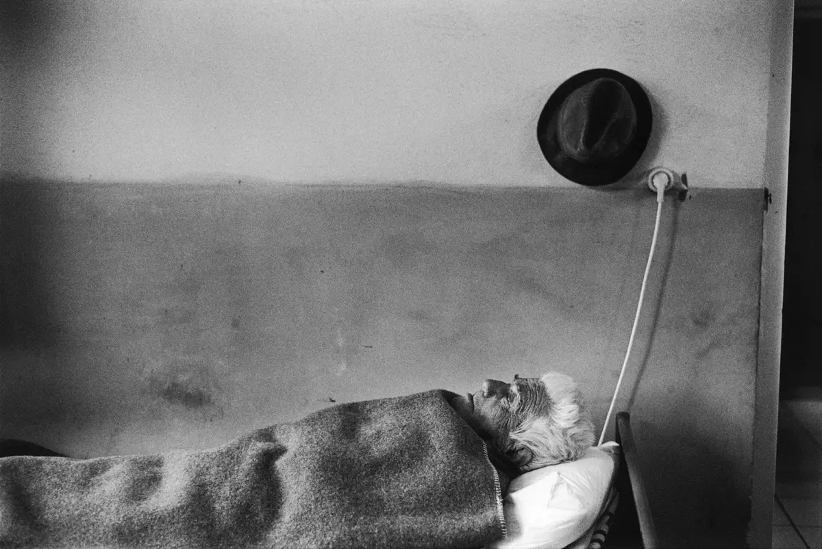 Older person lying in bed, covered by blanket, in a bare room; a black fedora hangs on the wall above an electrical outlet with a cord.