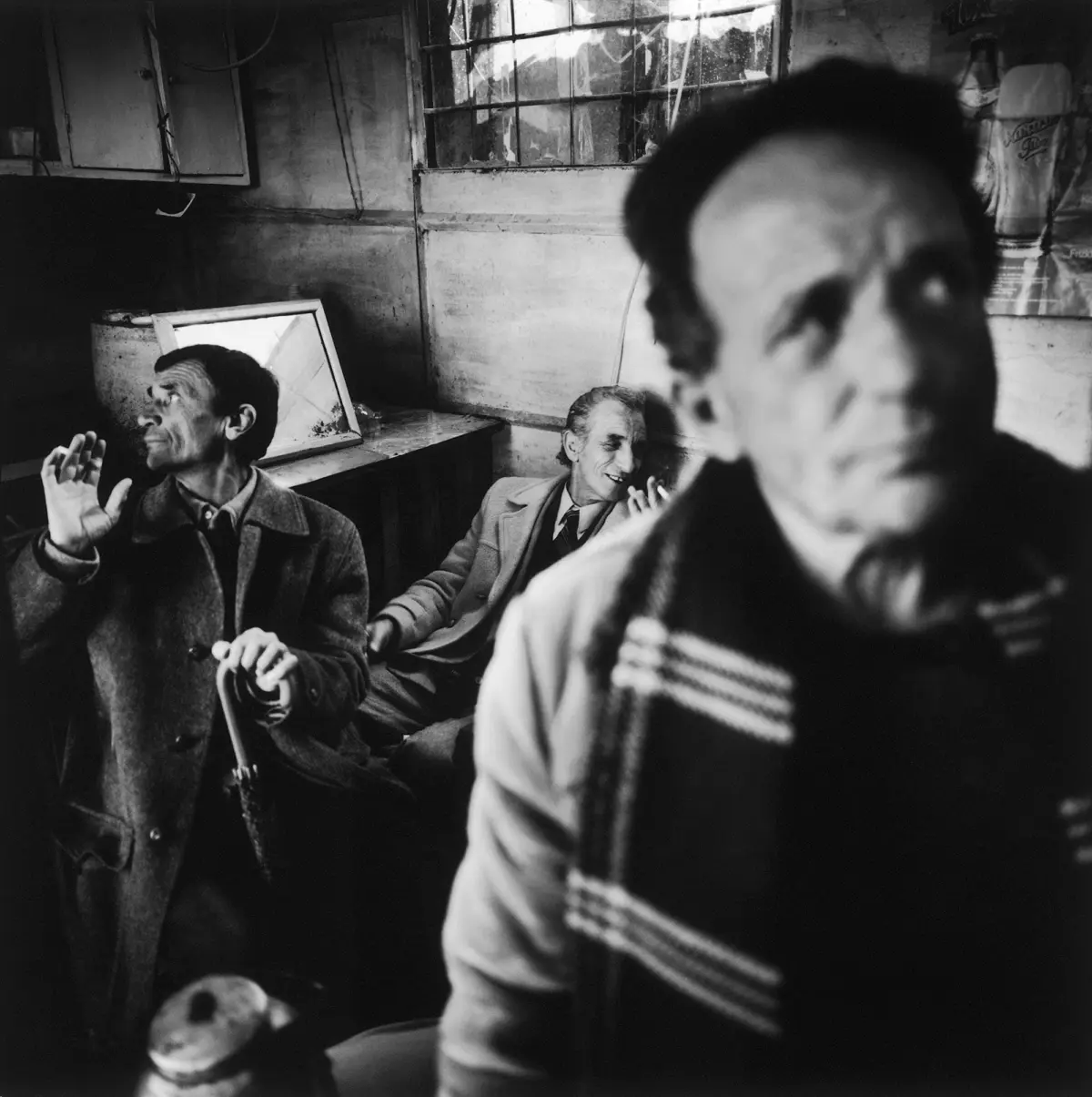Group of middle-aged men in a grimy, dim room; one man gestures with raised hands while others converse near a barred window.