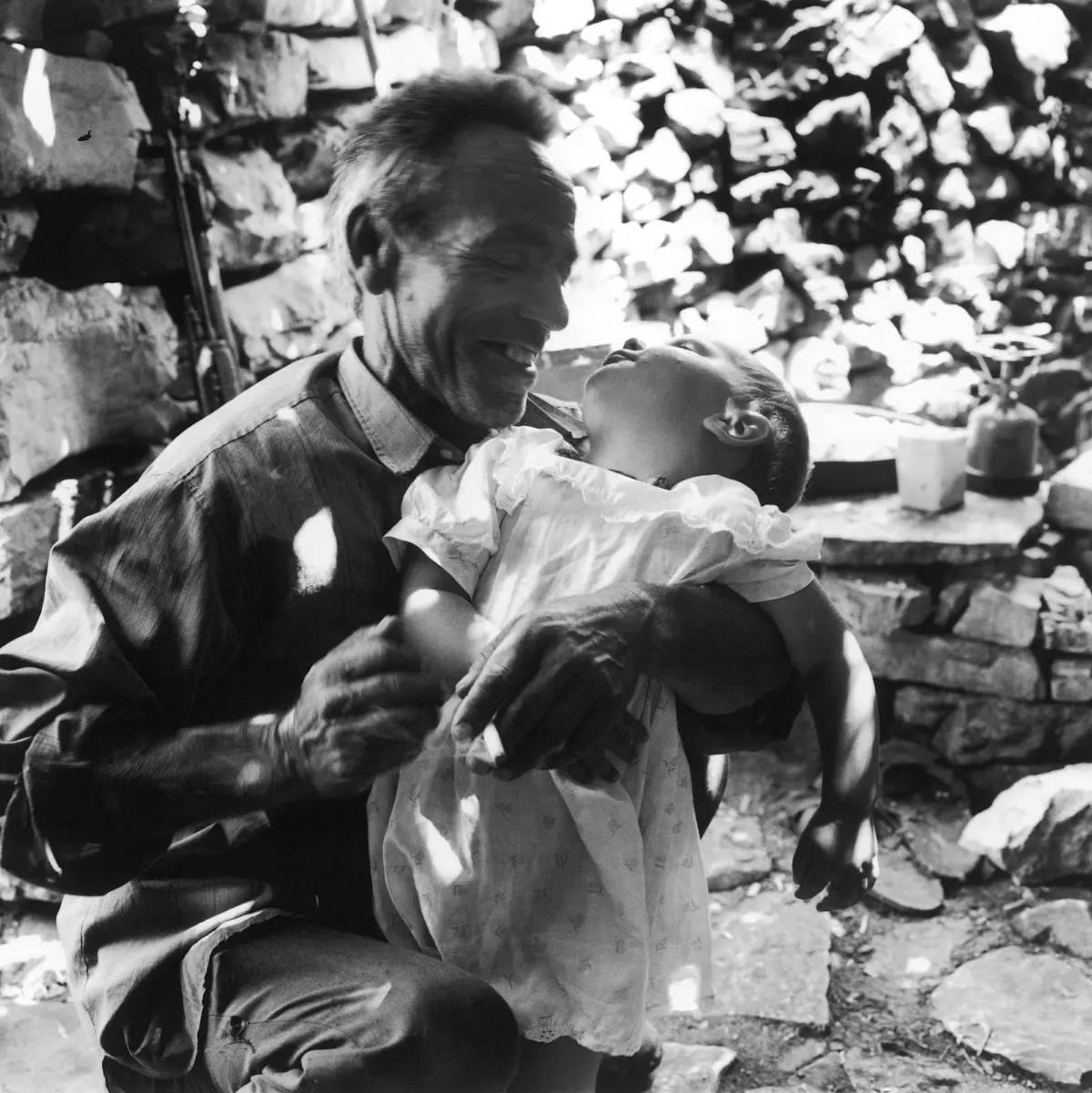 Adult embraces toddler in a lace dress; they smile outdoors beside a stacked stone wall.