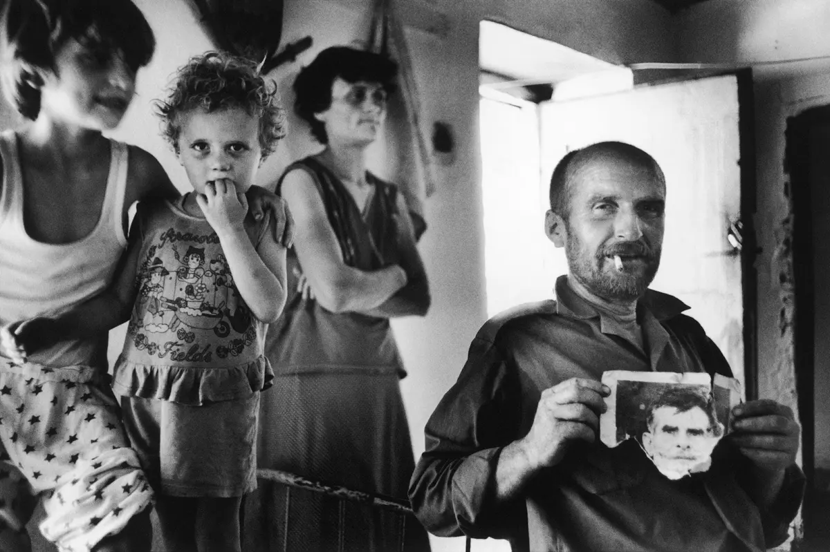 Adult man smoking while reading a magazine; curly-haired child beside him with fingers in mouth in an indoor setting.