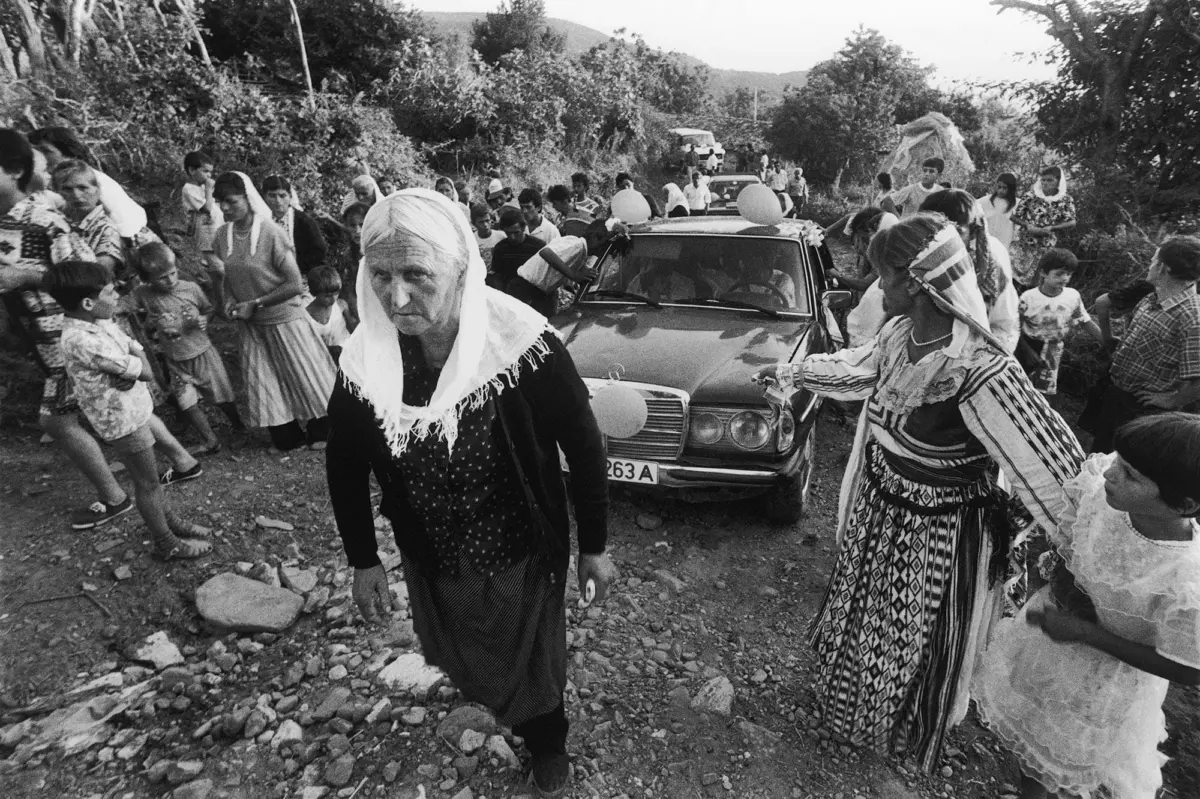 Elderly woman in a headscarf walks along a rocky rural road beside a decorated car with balloons as villagers gather.