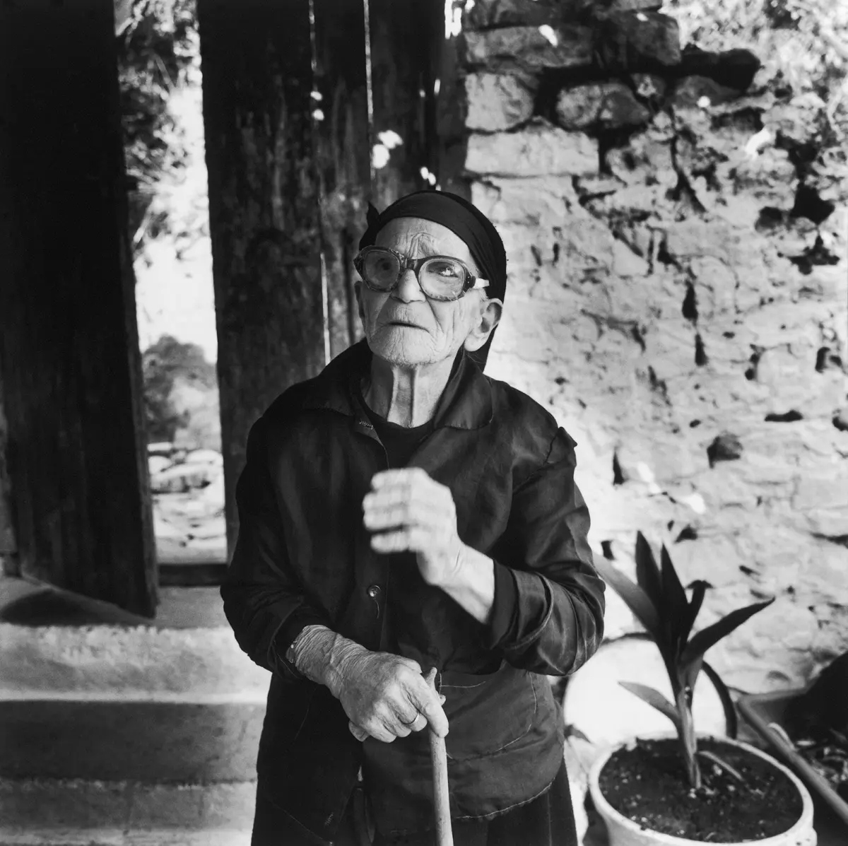 Elderly person wearing a headscarf and glasses, holding a wooden cane, standing beside a stone wall outdoors.