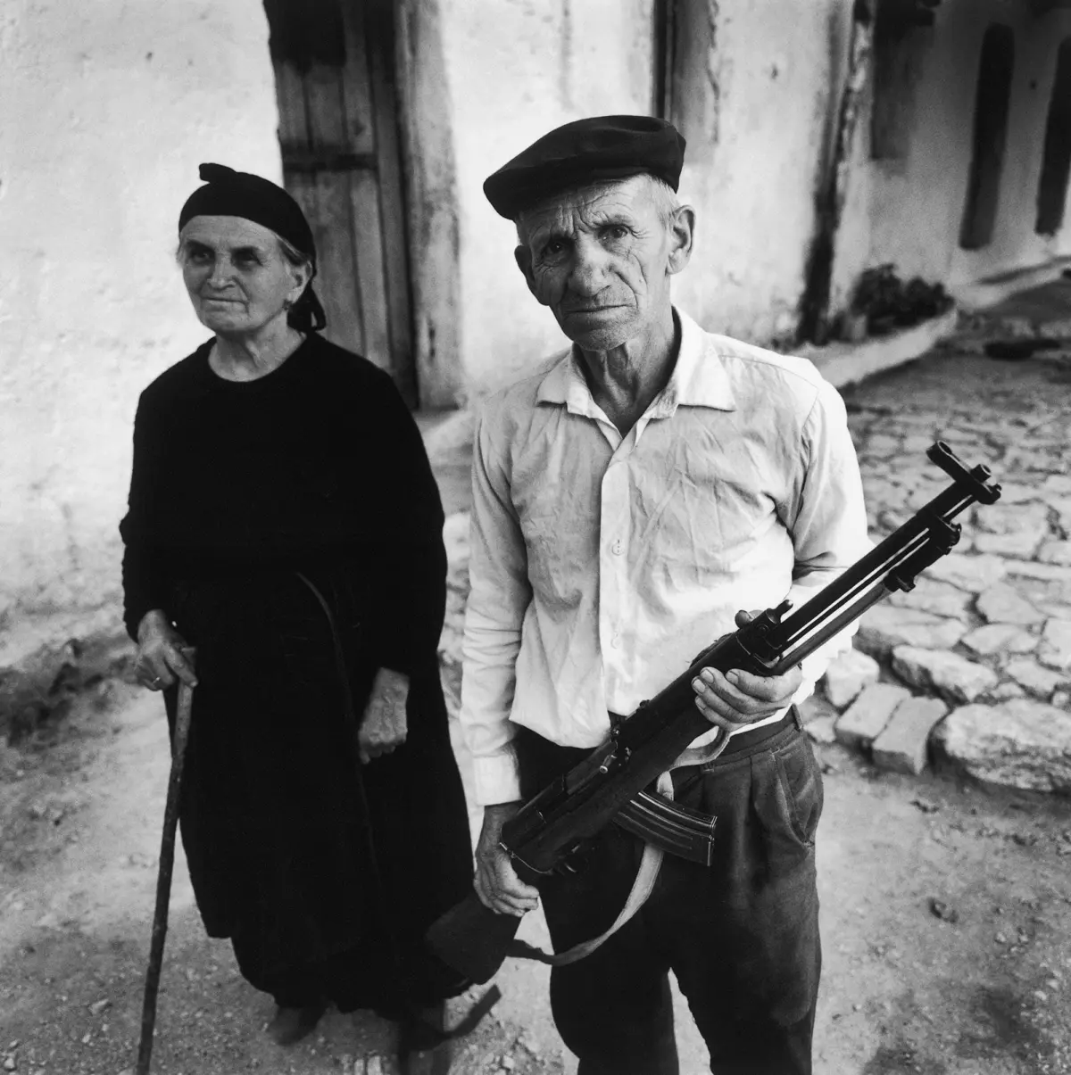 Older adult man holding a rifle stands on a cobbled village street beside an older adult with a cane; whitewashed building in background.