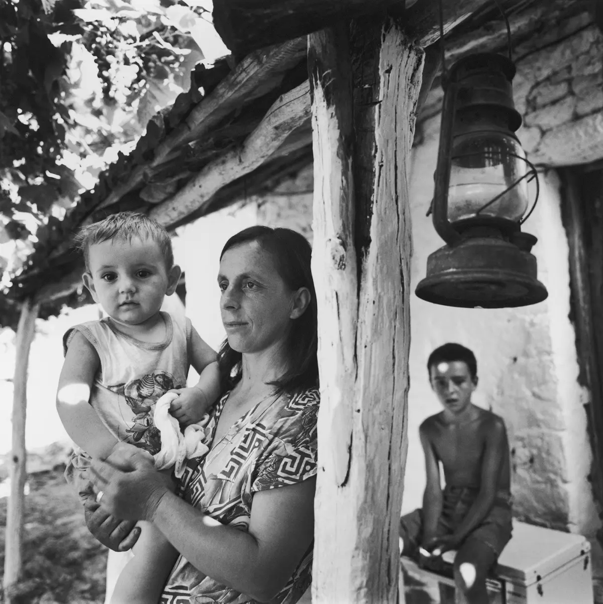 Woman holds toddler beside weathered wooden post with hanging lantern in a rustic outdoor setting.