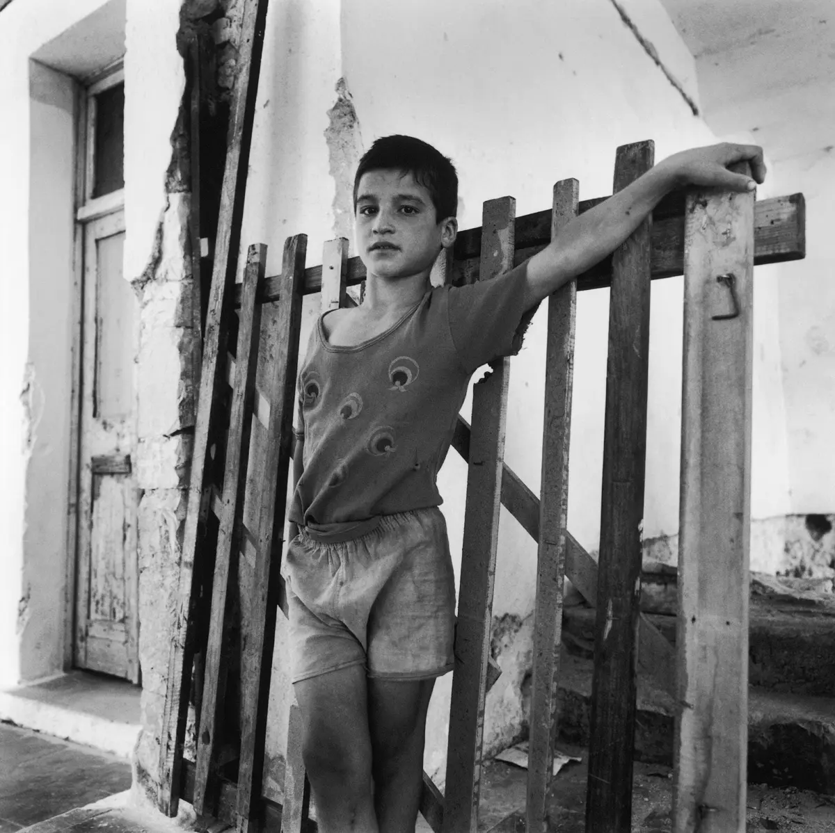 Boy leans on a weathered wooden fence outside a crumbling plaster building.