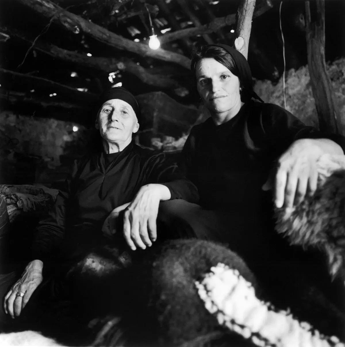 Two people in dark clothing and head coverings inside a dim cave, with a hanging light bulb; one hand reaches toward the camera.