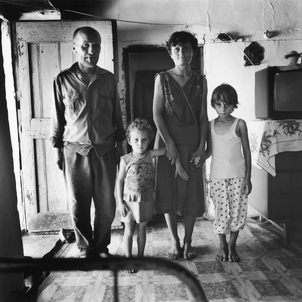 Adult man, adult woman, and two young girls stand together in a worn, rustic room with an old door and a television.