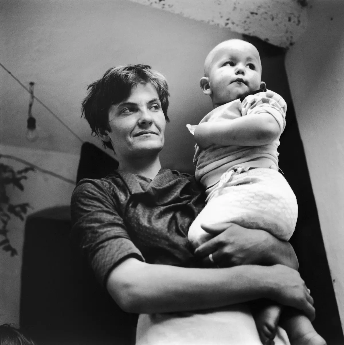 Adult with short hair holds infant on shoulder; infant wrapped in quilt, both looking upward indoors near a hanging bulb.