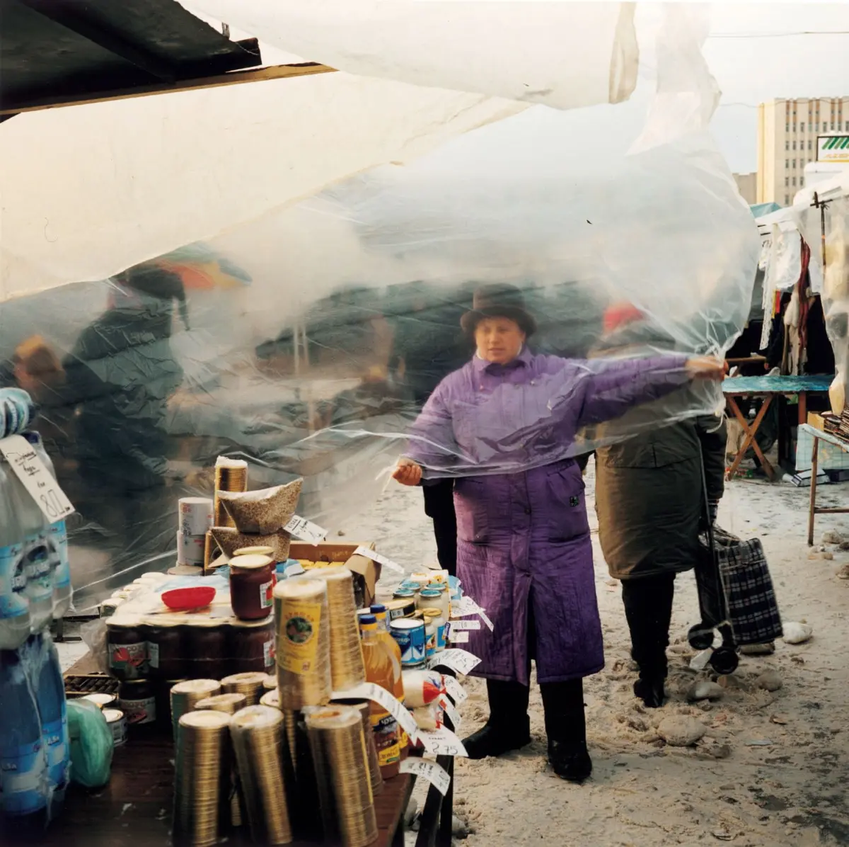 Woman in purple coat and black hat pulls back clear plastic sheeting at an outdoor market stall, with canned goods on display.