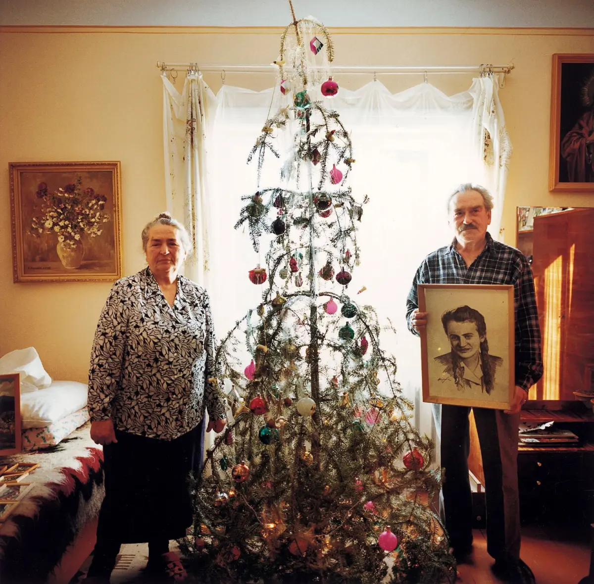 Elderly couple posing beside a decorated Christmas tree in a living room; man holding a framed portrait.