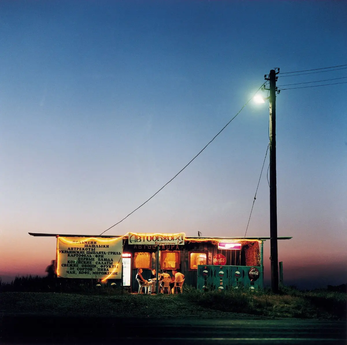 Roadside food stall at dusk with customers seated inside; banners with Cyrillic text, and a tall utility pole with a streetlight nearby.