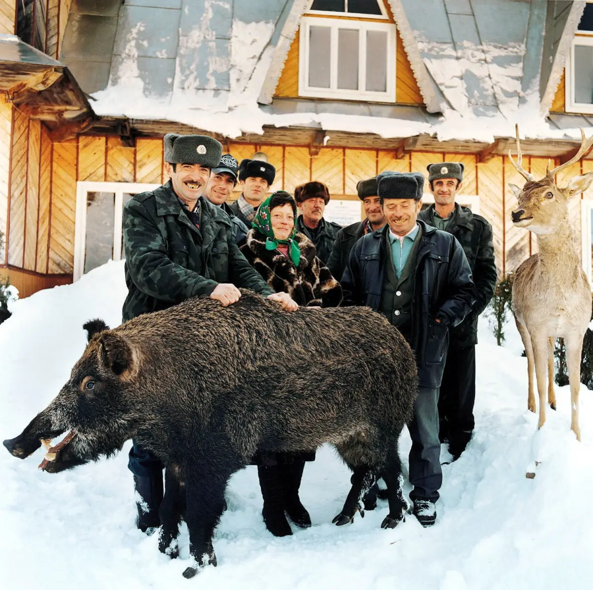 Group of people in fur hats posing with a large wild boar in a snowy yard in front of a wooden lodge; deer statue nearby.