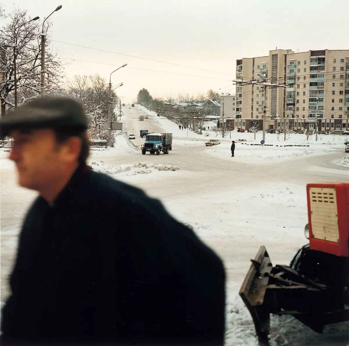 Blue dump truck drives along a snow-covered urban street; blurred man in foreground; residential buildings in the background.