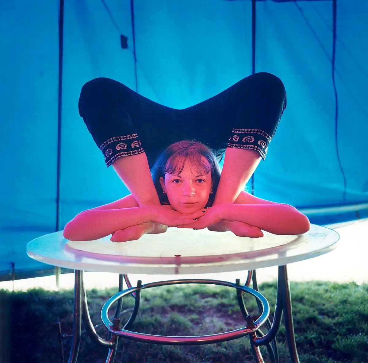 Contortionist performs a backbend over a round table outdoors under a blue tent.