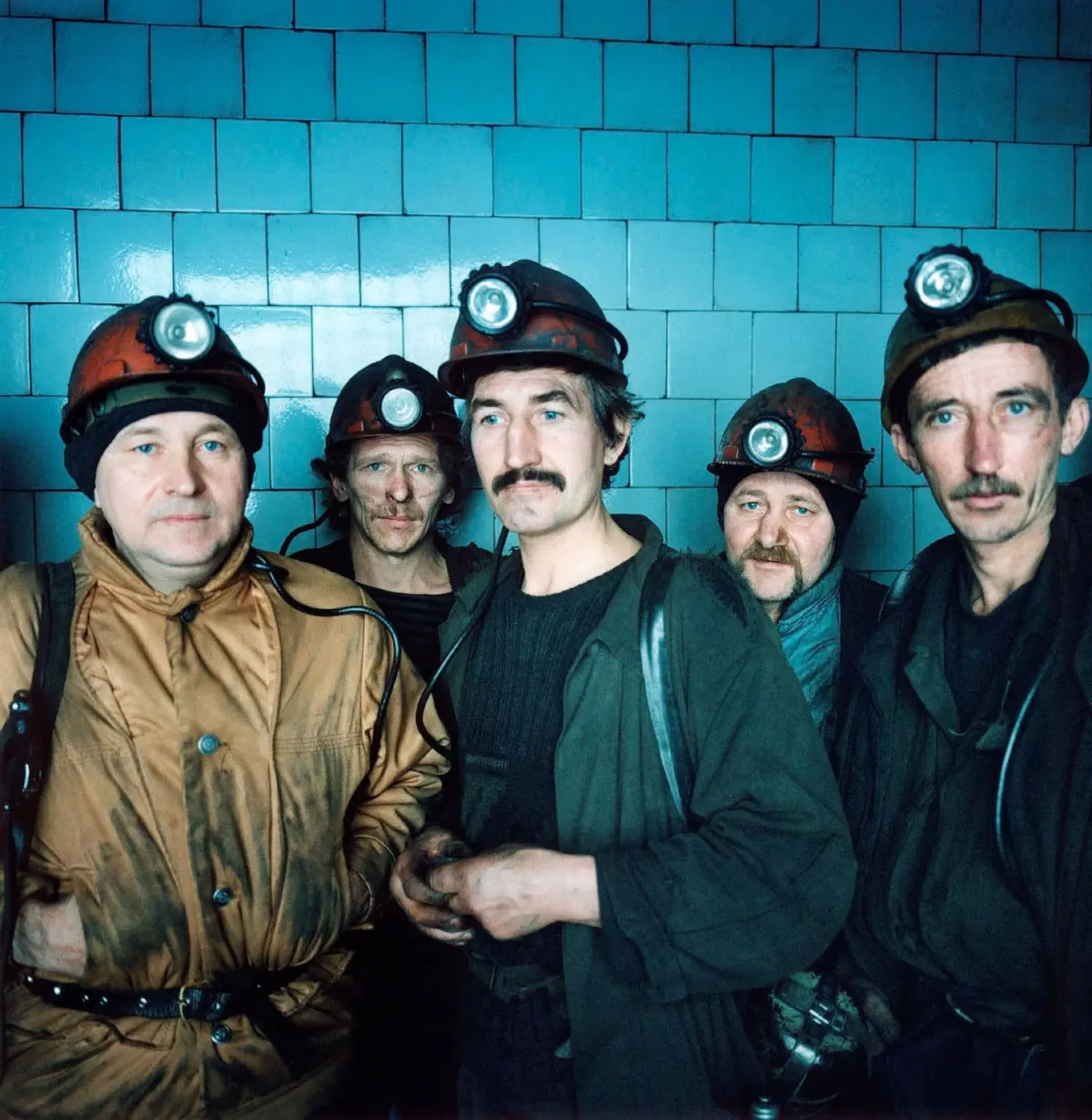 Five miners wearing headlamps and protective jackets pose together in front of a blue tiled wall.