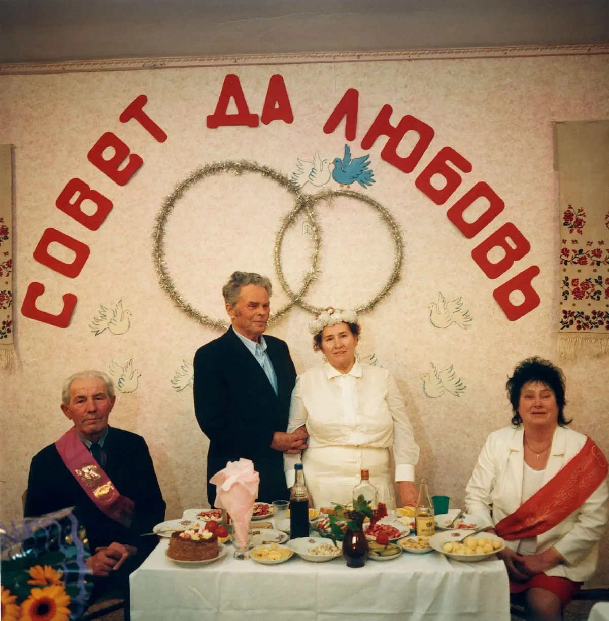 Bride and groom pose at a wedding reception with overlapping ring decorations behind; guests sit at a food-filled table