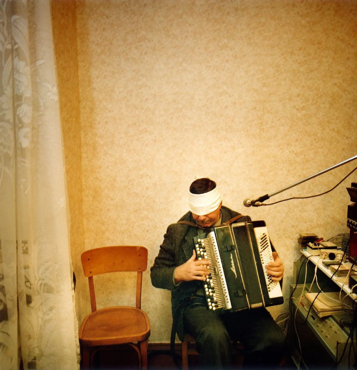 Musician with white head wrap plays accordion in a home studio with beige wallpaper, microphone, and cables.