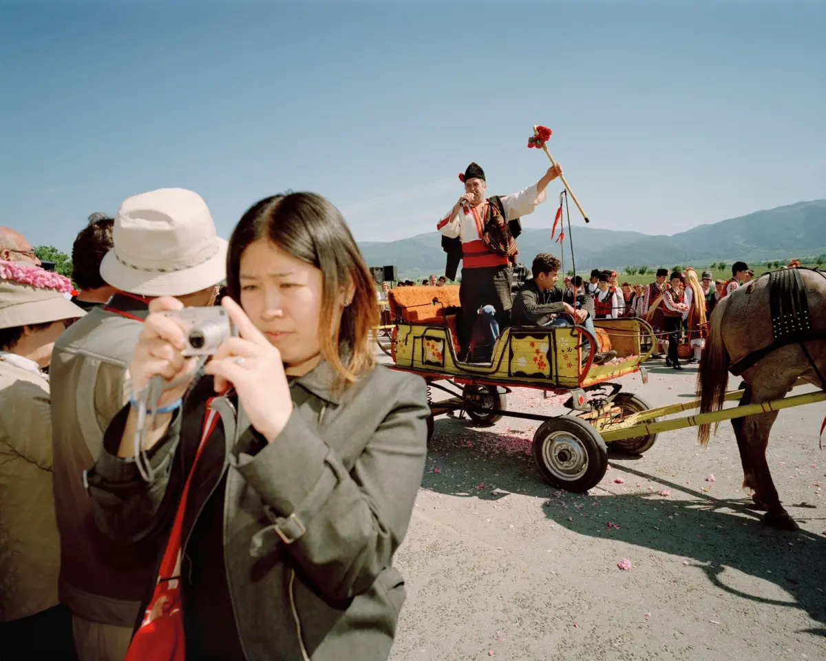 Performer in traditional attire on a decorated horse-drawn carriage, speaking into a microphone and waving a red-flower staff at an outdoor festival.