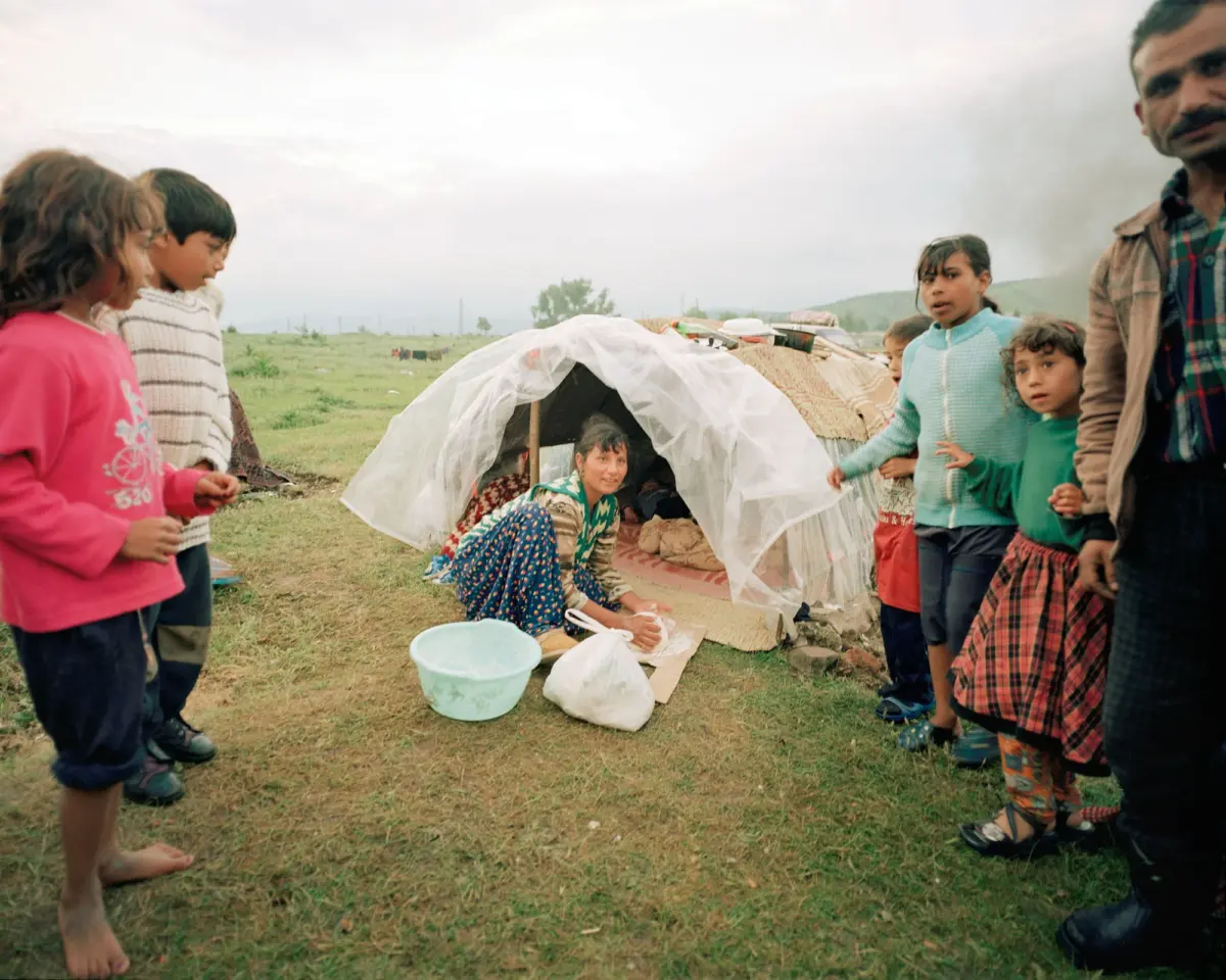Group of children and an adult standing around a makeshift shelter covered with plastic sheeting in a grassy field.