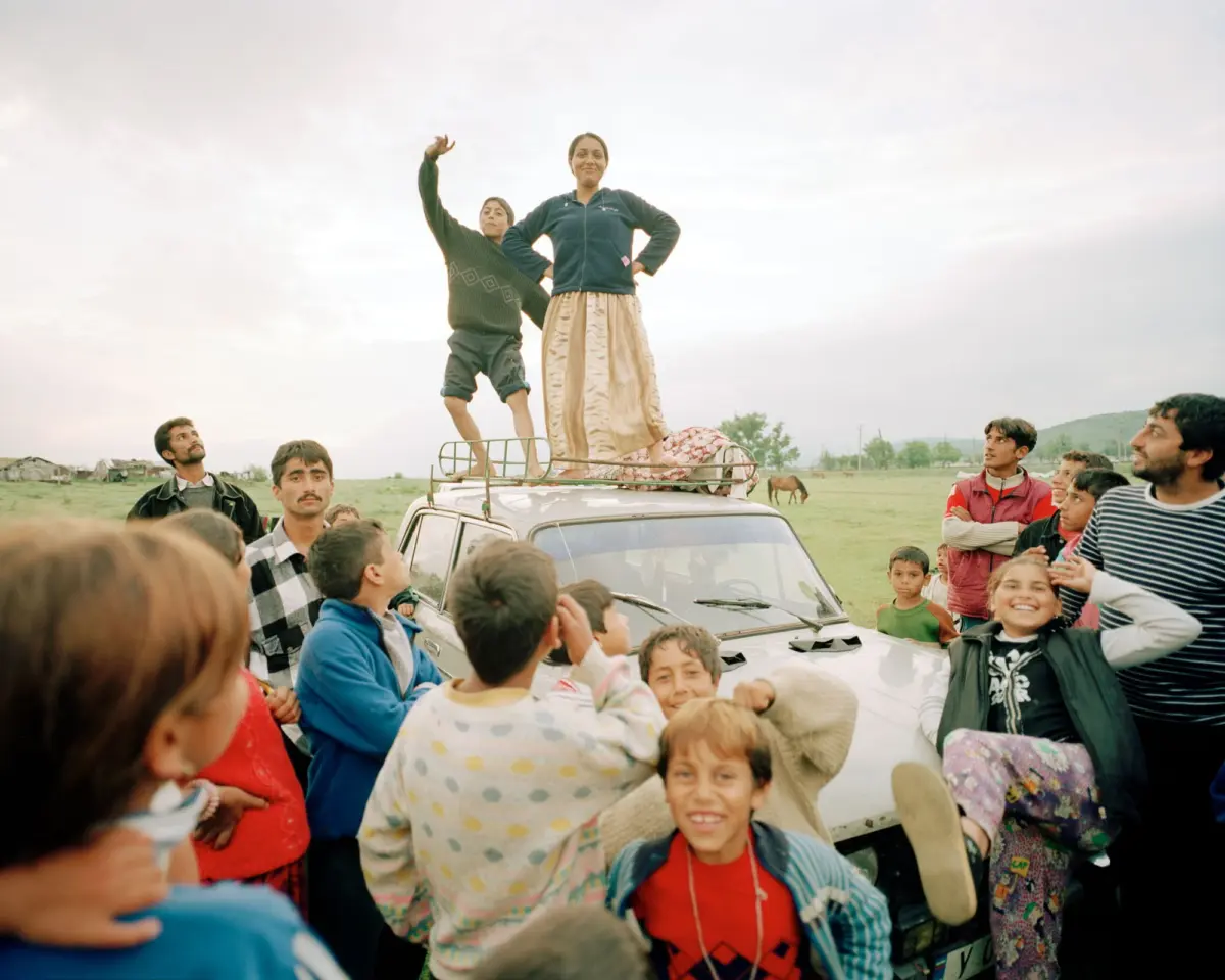 Two people stand on a car roof, posing with arms raised, while a crowd of children gathers in an open field.