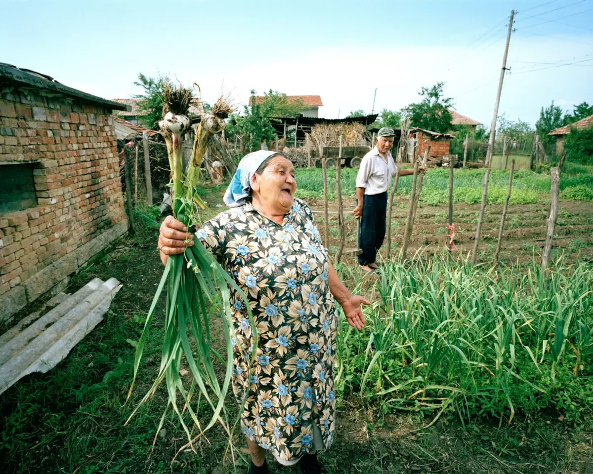 Elderly woman in floral dress holds green onions in a rural garden; man in cap stands by a wooden fence, brick buildings in background.