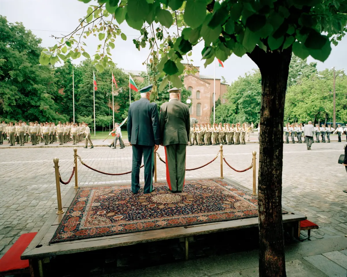 Two military officers in dress uniforms stand on a carpeted platform, facing away, watching a line of soldiers in a flag-lined courtyard.