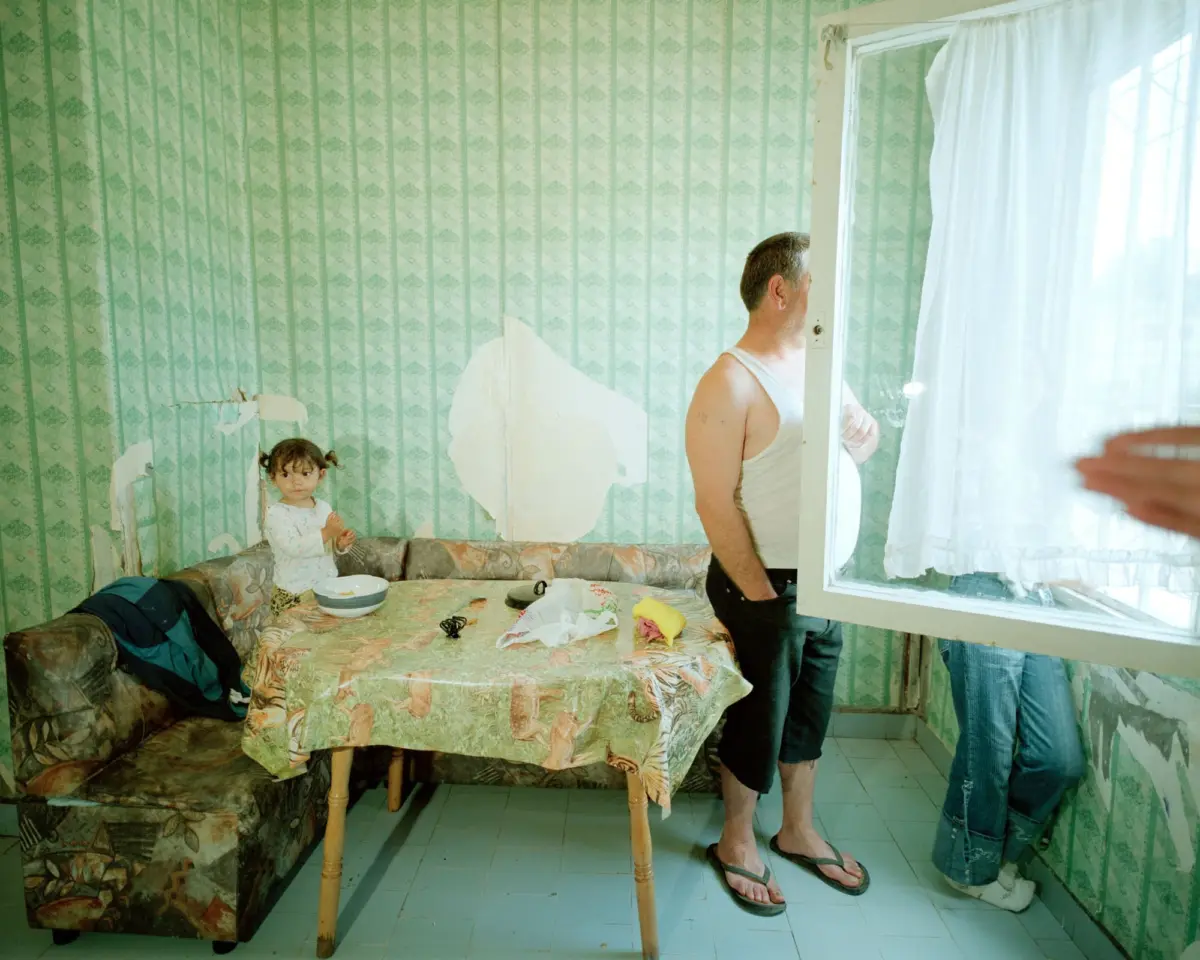Adult in sleeveless shirt stands by window, looking out, in cluttered living room with peeling green wallpaper and floral sofa, a child nearby.