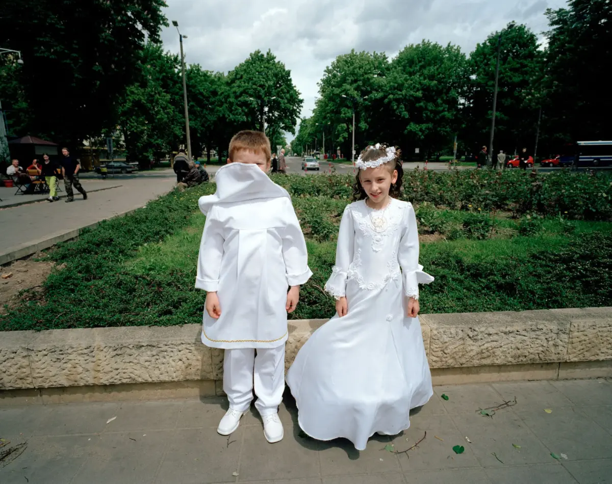 Two children in white ceremonial outfits stand by a stone wall in a park; one child faces camera with a floral headband, the other has back to camera.
