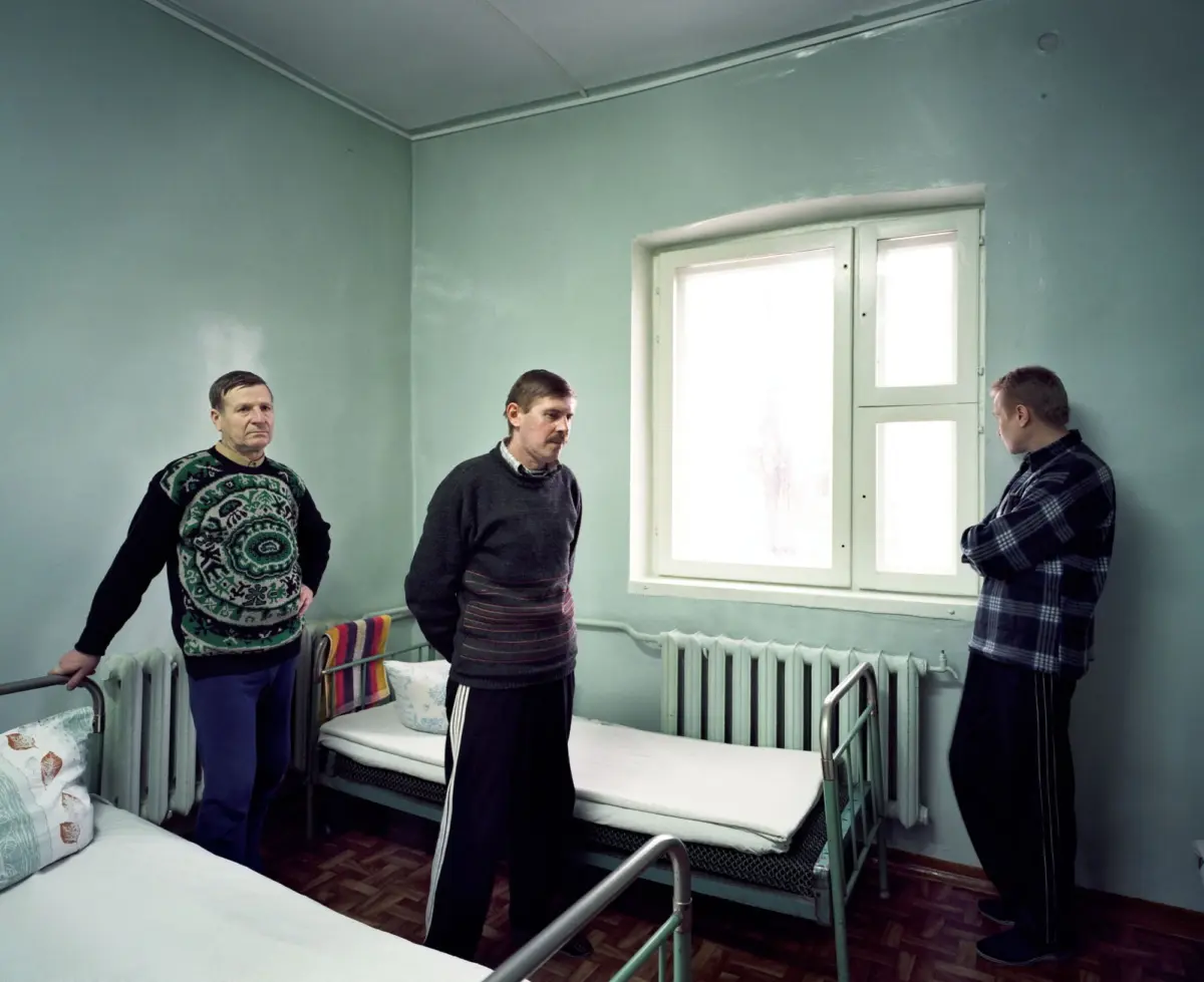 Three adults stand by metal hospital beds in a pale-green ward near a large window.