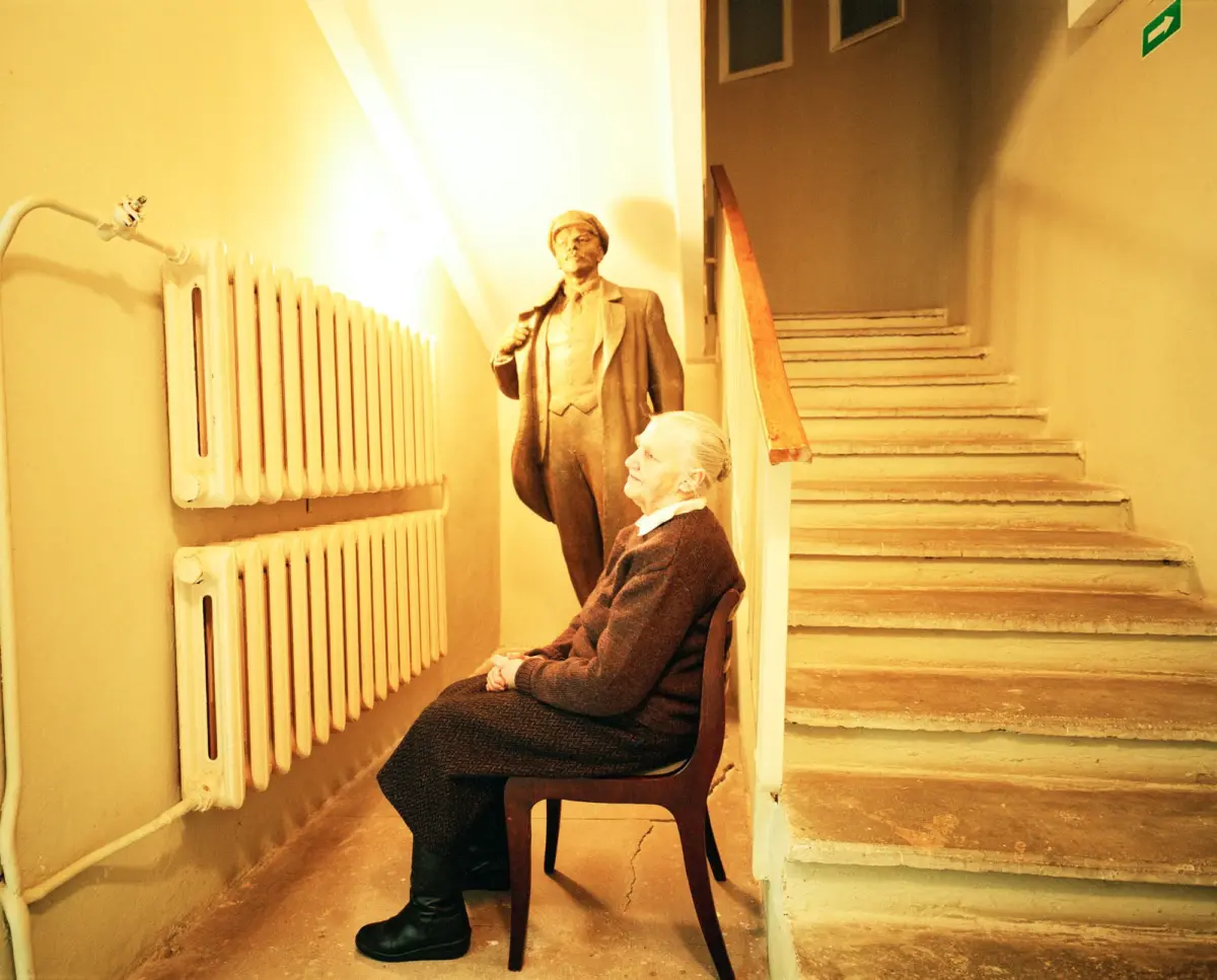 Elderly woman seated on a chair in a yellow hallway beside a suit-clad statue; radiator on the left and a staircase on the right.
