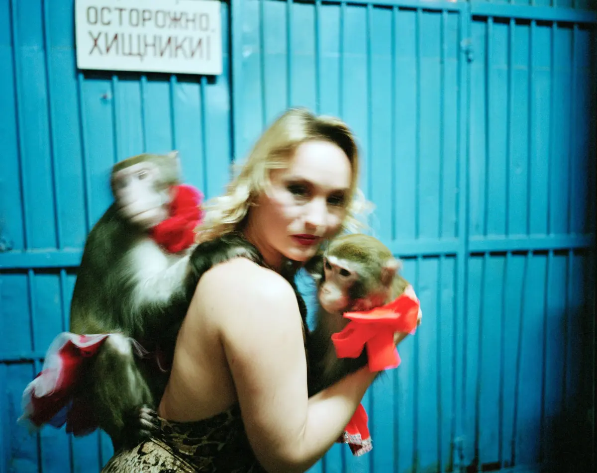 Blonde woman holding a toddler, with a dog wearing a red scarf beside her, in front of a blue metal fence.