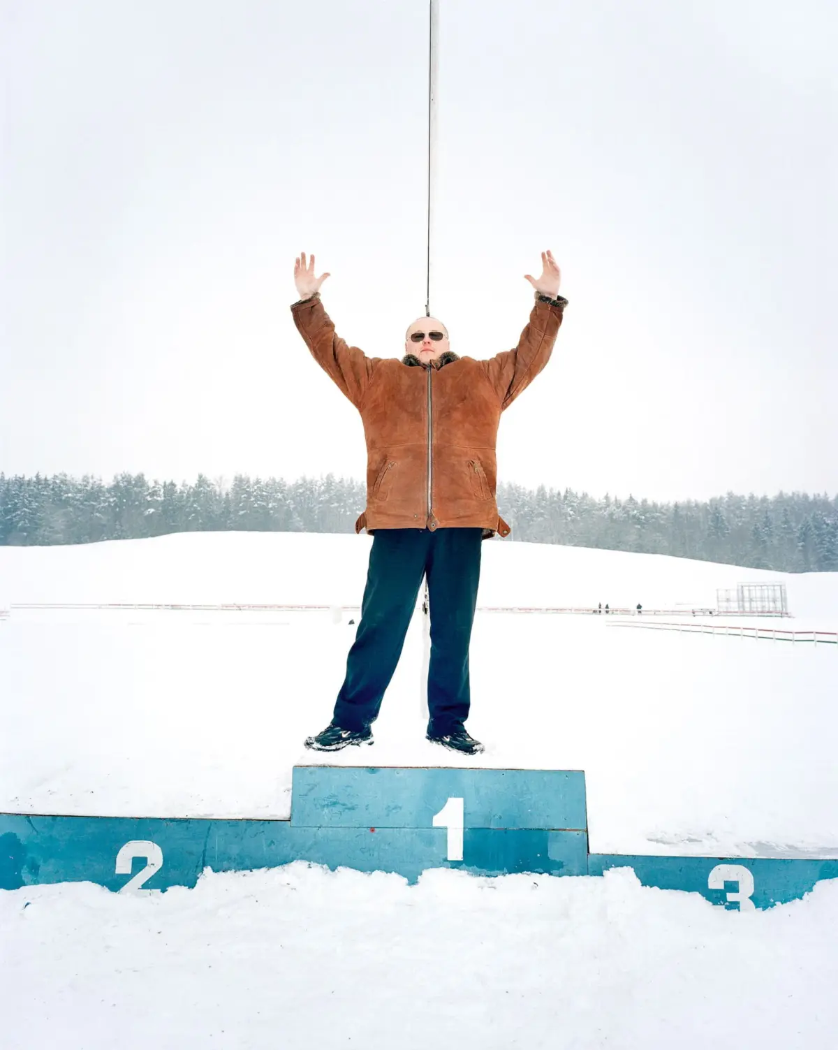 Person on snow-covered ground raises arms atop a blue podium marked 1, wearing brown shearling jacket and sunglasses; rope overhead.
