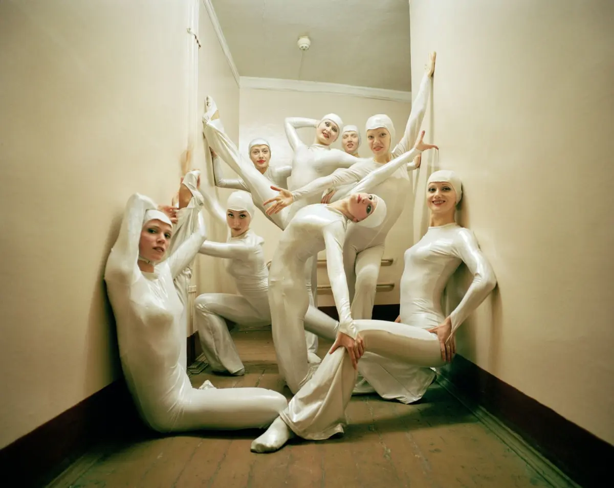 Group of performers in white latex bodysuits and caps posing dramatically in a narrow stairwell hallway.