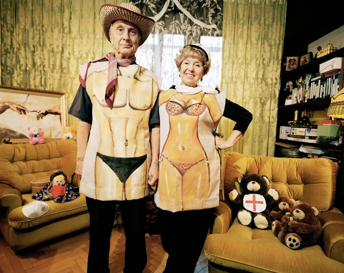 Two seniors wear torso-shaped aprons, posing and smiling in a living room with green patterned curtains, a yellow sofa, and stuffed bears.