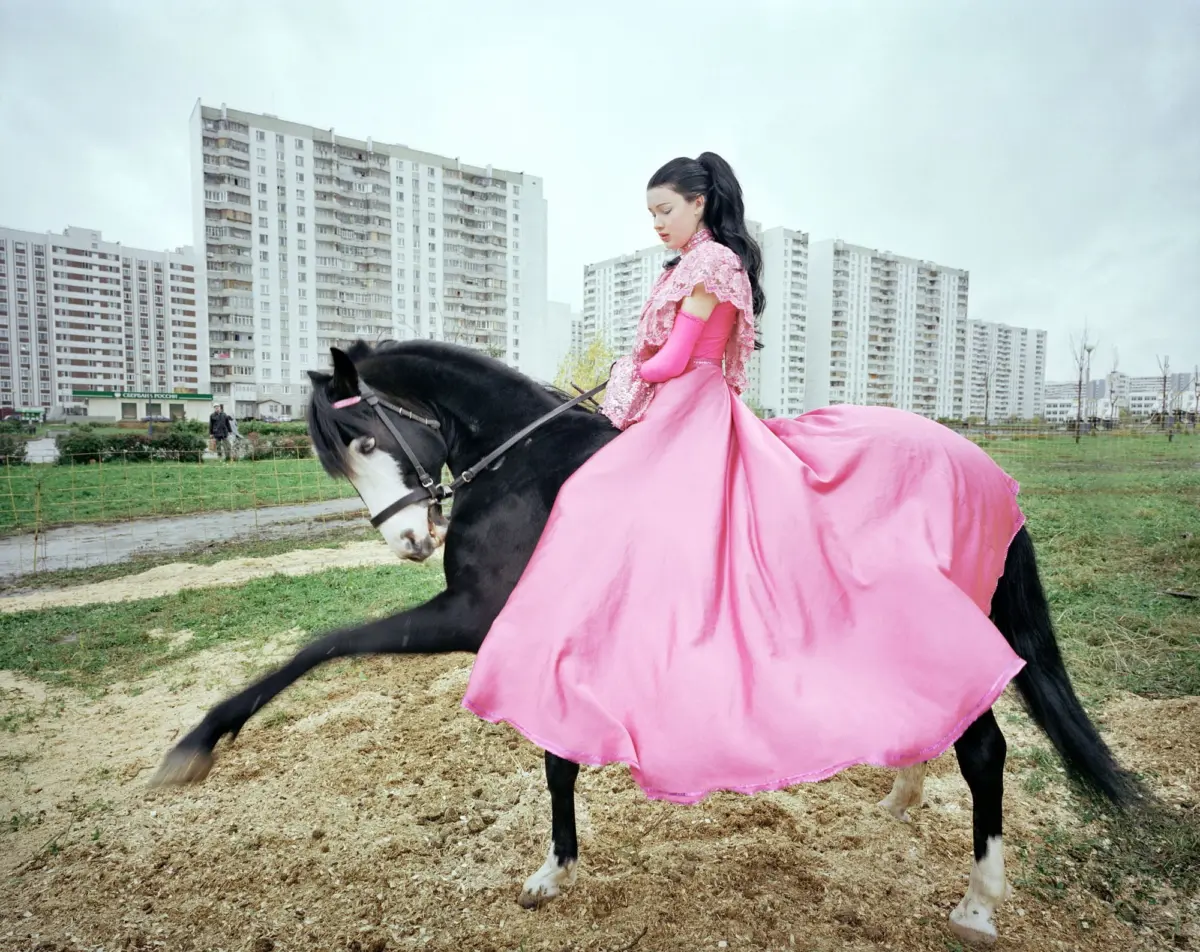Woman in a pink gown rides a black horse with pink cape through a dirt lot, with tall apartment buildings in the background.