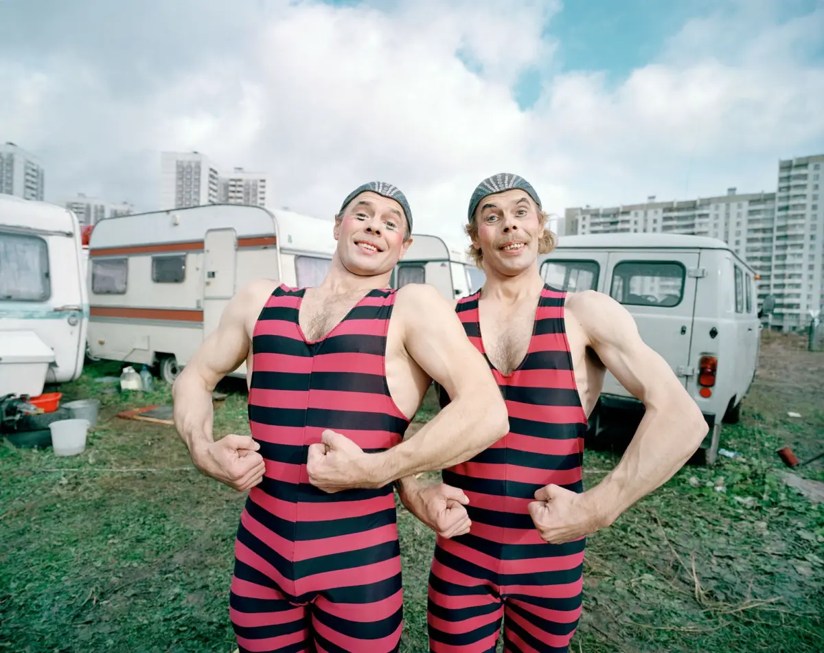 Two men in pink-and-black striped swimsuits with swim caps flex their biceps at a caravan park, with caravans and apartment buildings in the background.