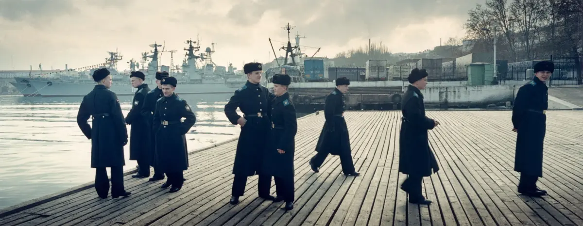 Group of sailors in dark uniforms and fur hats stand on a wooden dock by a harbor with warships in the background.