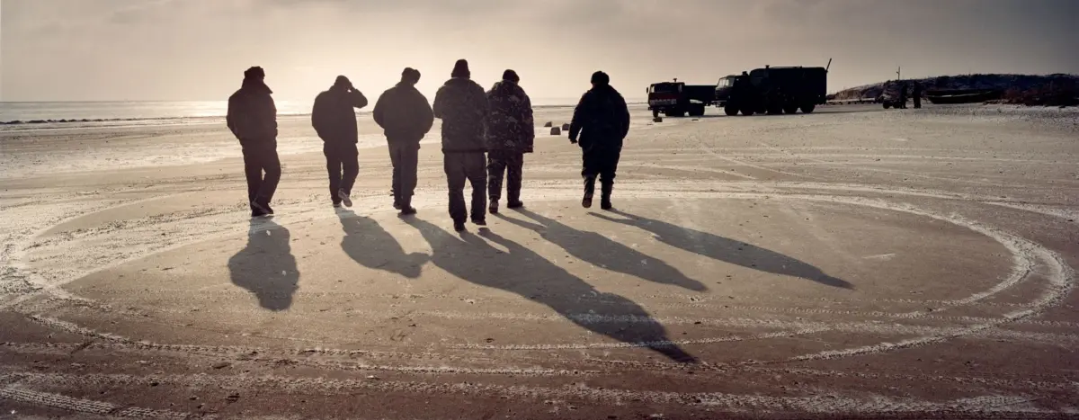 Group of soldiers walking along a sandy beach at sunset, casting long shadows; trucks parked near the dunes in the distance.