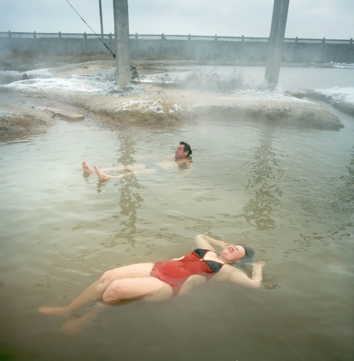 Person in red swimsuit floating on back in muddy, steaming pool, with concrete columns and railing in the background.