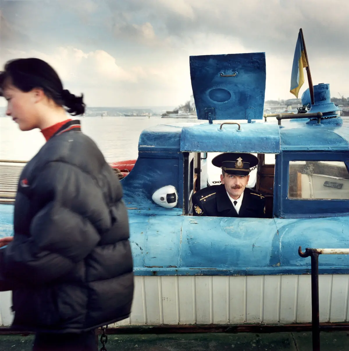 Naval officer in uniform peeks from a blue boat cabin; blurred passerby in foreground, harbor and distant city in background.