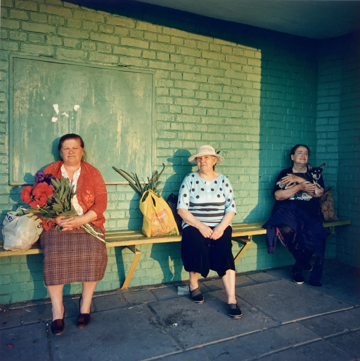 Three women pose against a teal brick wall; left holds red bouquet, middle in polka-dot blouse, right cradles small dog.