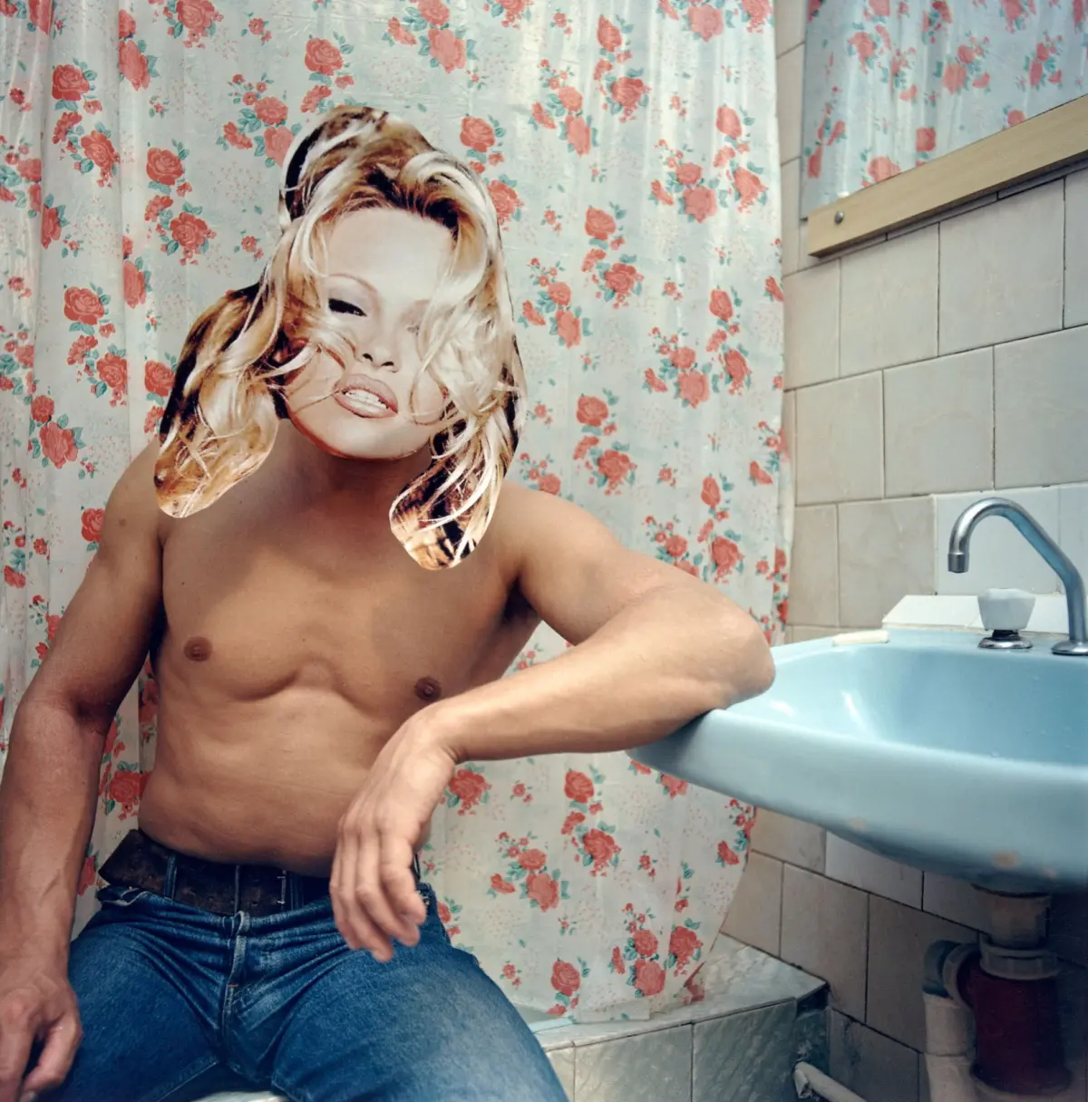 Shirtless adult person with blond hair sits by a pale blue bathtub in a bathroom, floral pink curtain behind, wearing a cutout face mask.
