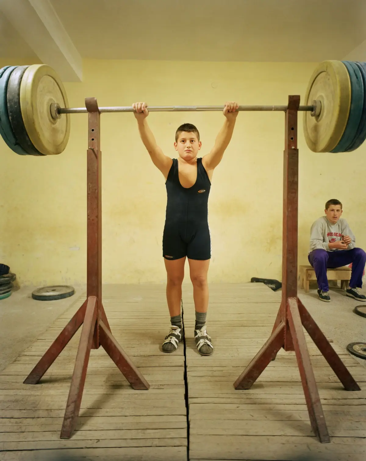 Young weightlifter in a navy singlet overhead-presses a loaded barbell on a weightlifting platform in a gym.