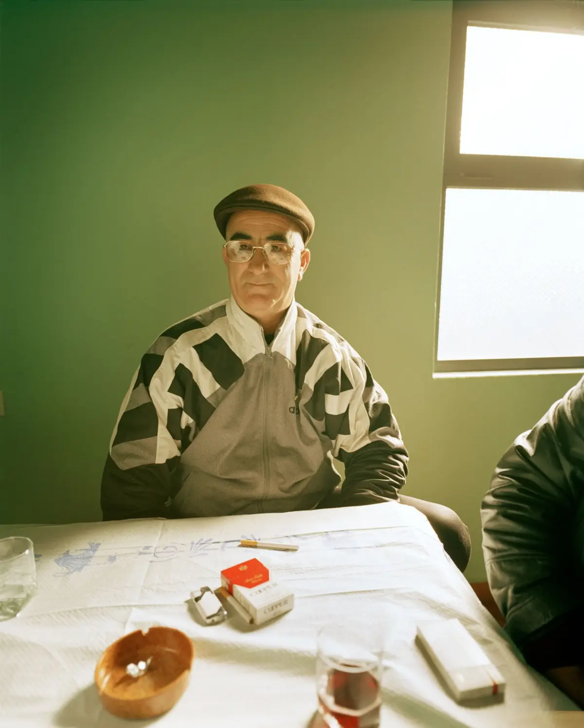 Older person in brown flat cap and diamond-patterned jacket sits at a table by a green wall, looking at the camera, near a bright window.