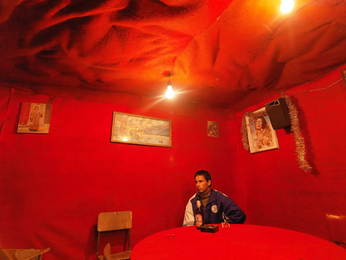 Person in blue jacket sits at a red round table in a red-walled room, holding a beer bottle; hanging bulbs and framed art decorate the space.