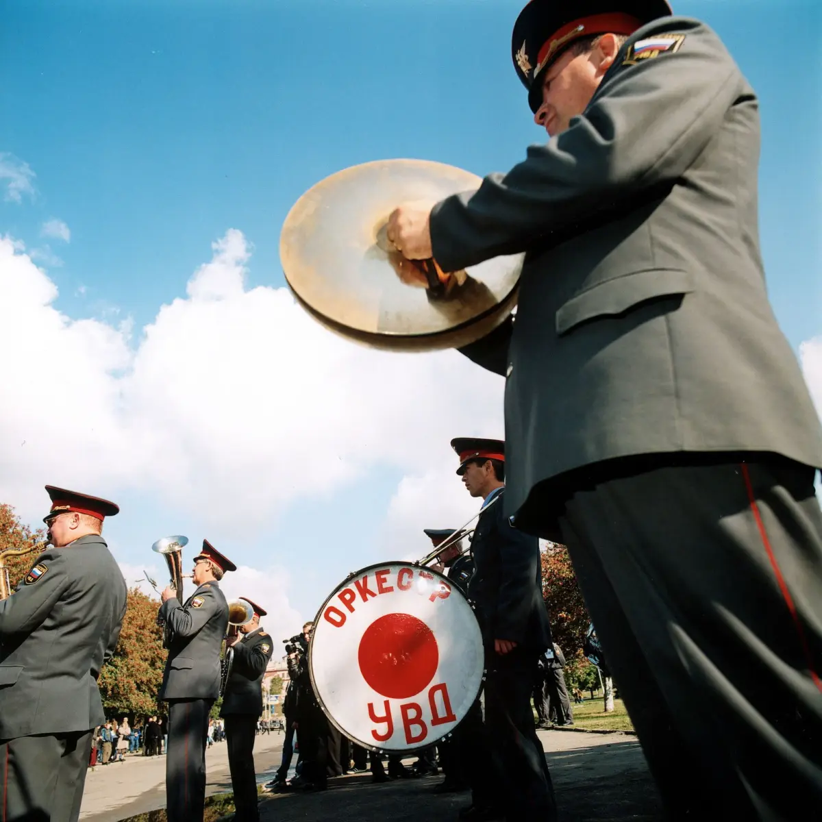 Military band members in gray uniforms with red-trimmed peaked caps playing large bass drums during an outdoor parade.